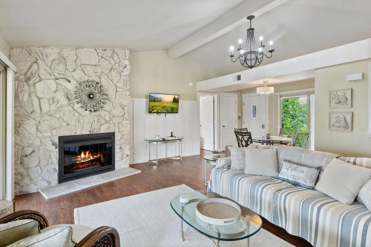The image shows a warm, inviting living room with a stunning stone fireplace as the centerpiece, surrounded by a comfortable sofa and stylish coffee table under a glowing chandelier. The space is designed for relaxation and gathering, ideal for quiet mornings with coffee or lively evenings with family and friends.