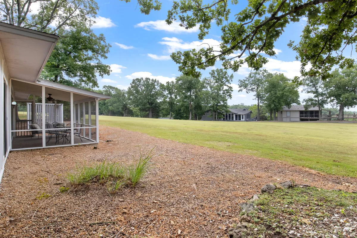 A peaceful screened porch overlooks an expansive green lawn, with a canopy of mature trees and the distant outline of community buildings visible through the foliage. The porch is inviting, set up for relaxed outdoor lounging, and is perfectly positioned to enjoy the view and tranquil atmosphere.