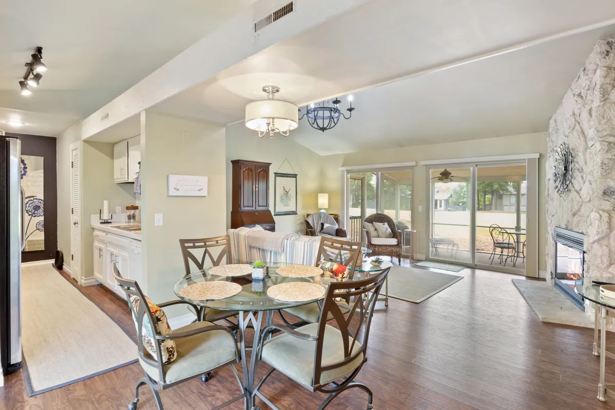 A cozy, sunlit open-concept living and dining space with a glass dining table, stone fireplace, large windows, and comfortable seating. The room is arranged for socializing, dining, and relaxation, capturing the inviting spirit of the home.