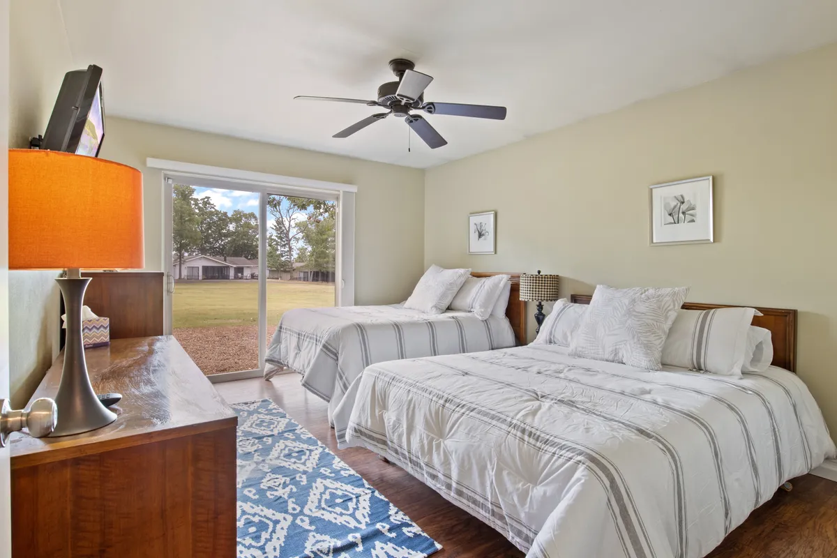 A double queen bedroom with two beds dressed in crisp white bedding, a decorative lamp, and a sliding glass door that leads to a scenic outdoor view. The room is bathed in soft natural light, making it ideal for families or friends seeking comfort and a peaceful start to their day.