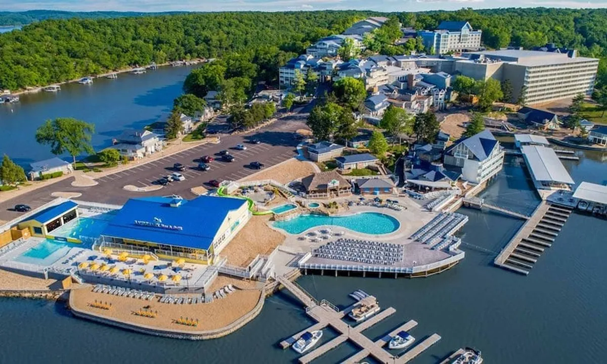 A vibrant outdoor pool scene featuring blue water, sun loungers, boats, docks, and a nearby restaurant area. Guests can be seen swimming, socializing poolside, and preparing for boating adventures, all set against a lively, resort-like backdrop.