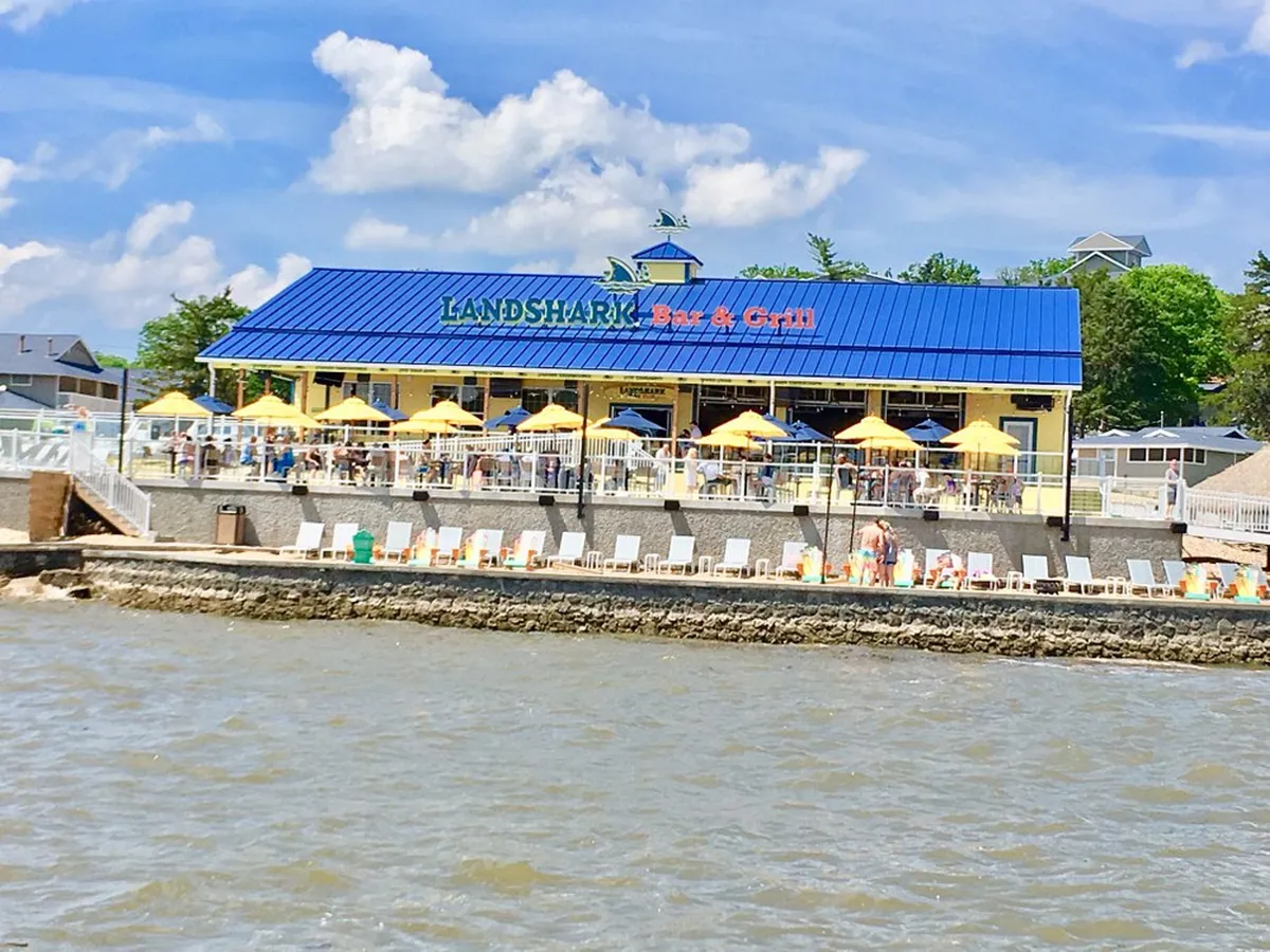 The Landshark Bar & Grill’s colorful building sits beside the water, with yellow umbrellas shading outdoor tables on a lively waterfront patio. Guests enjoy food and drinks, and the festive mood hints at sunset views and lakeside dining experiences.