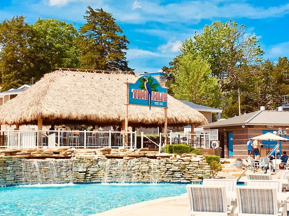 A lively outdoor pool scene featuring a Tiki Hut bar, cascading waterfalls, and plenty of comfortable seating surrounded by lush greenery. Guests are seen socializing at the bar, relaxing in the sun, and enjoying the resort-style atmosphere.